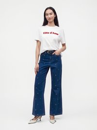 Γυναίκα φορώντας ένα λευκό T-shirt "Côte d'Azur", μπλε φαρδιά τζιν με μικρά λευκά σχέδια, μαύρη ζώνη και λευκά παπούτσια με χαμηλό τακούνι.