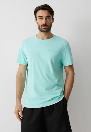 Mężczyzna o ciemnych włosach i brodzie, ubrany w jasnoniebieski t-shirt i czarne spodnie, stojący na tle jednolitego jasnoszarego tła.