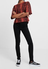 Osoba stojící se zkříženýma rukama, oblečená v krátkém topu s potiskem červeného hada, černých skinny kalhotách a černo-bílých teniskách.