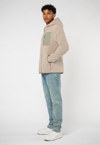 Fleece-Hoodie in hellbeige mit einer grünen Brusttasche, kombiniert mit hellblauen Jeans und weißen Sneakers. Verfügt über eine Kapuze mit Kordelzug.