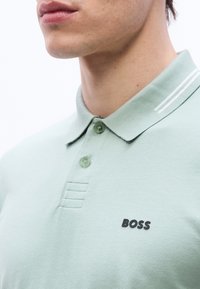 Κοντινό πλάνο ενός ατόμου που φοράει μια ανοιχτοπράσινη μπλούζα πόλο με δύο κουμπιά και το  λογότυπο "BOSS" στο στήθος.