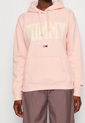 Osoba ubrana w jasnoróżową bluzę z kapturem Tommy Jeans z przednią kieszenią oraz brązowe spodnie, pokazana tylko od tułowia i ramion.