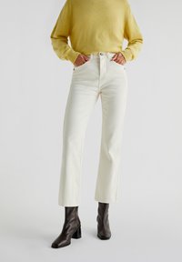 Witte high-waisted jeans met een rechte pijp, gecombineerd met een gele trui. Voorzien van zijzakken en zichtbare stiksels. Bruine hakboots.