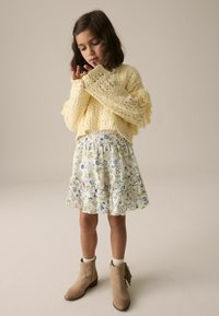 Hellgelbe Strickjacke mit fransigen Ärmeln, kombiniert mit einem weißen Rock mit Blumenmuster und Rüschen, sowie tanfarbenen Ankle Boots mit Verzierungen.