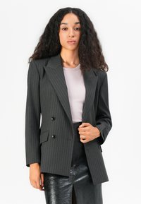 Blazer nero con righe contrastanti e design doppiopetto, con rever a punta e due tasche frontali; indossato su un top lilla chiaro e una gonna nera lucida.