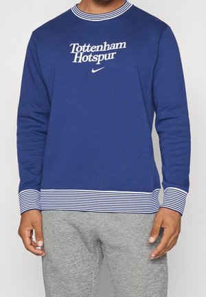 Mężczyzna ubrany w niebieską koszulę z długim rękawem z tekstem "Tottenham Hotspur" i logo Nike, w połączeniu z szarymi spodniami.