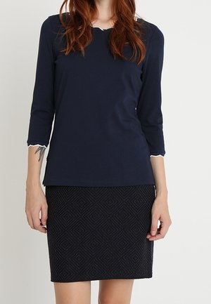Femme portant un haut bleu marine à manches trois-quarts avec bords festonnés et une jupe noire texturée longueur genou.