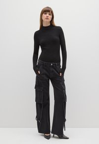 Schwarzes, eng anliegendes Rollkragenoberteil mit Schimmer; schwarze Cargo-Hose mit mehreren Taschen und weitem Bein; spitze schwarze Absätze mit silbernen Nieten.