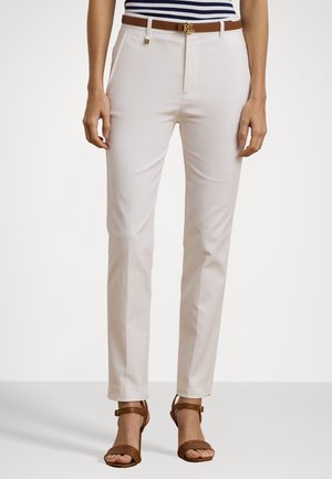 Femme portant un pantalon blanc slim avec une ceinture marron et des talons ouverts marron, debout devant un fond clair uni.