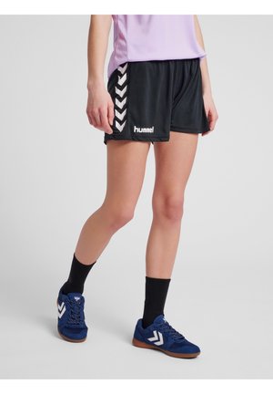 Person trägt schwarze Hummel-Sportshorts, schwarze Socken und blaue Sneakers mit weißem Chevron-Logo, steht auf einem hellgrauen Boden.