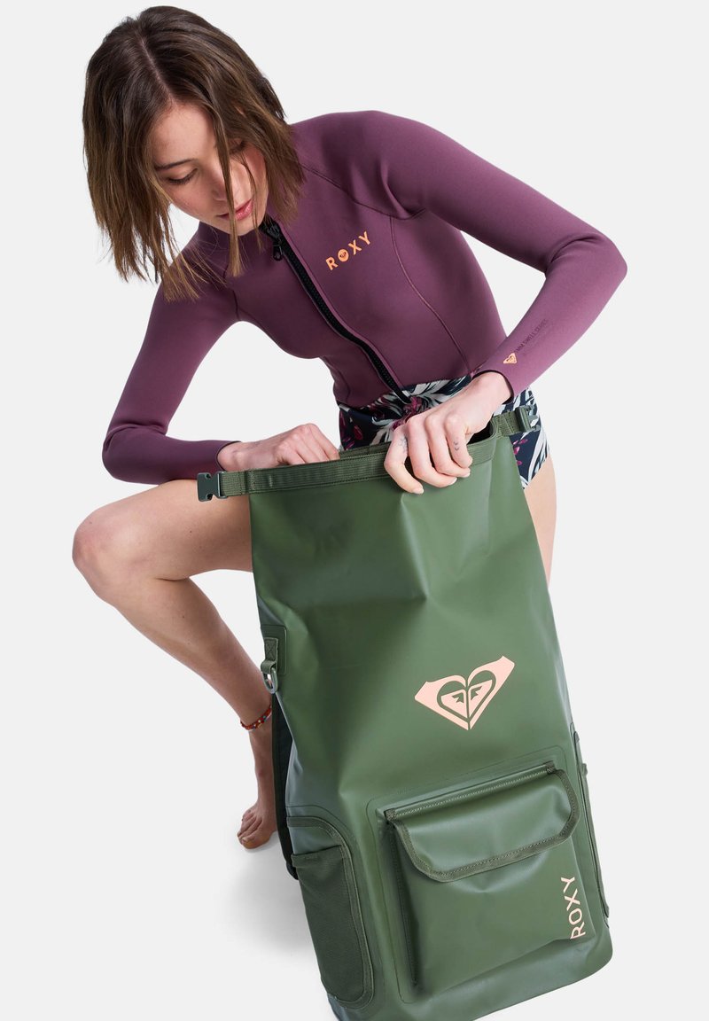 Sac à dos imperméable vert avec une grande ouverture, logo Roxy et une poche avant. L'utilisateur porte une combinaison de surf violette et ouvre le sac.
