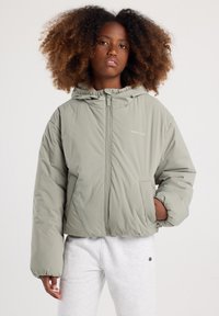 Giacca imbottita verde chiaro con cappuccio, chiusura frontale e tasche laterali; presenta una silhouette squadrata e polsini elastici.