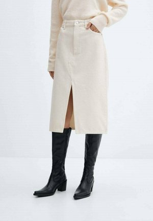 Femme portant une jupe midi crème avec une fente devant, un pull beige et des bottes noires montantes à talons, debout devant un fond uni.
