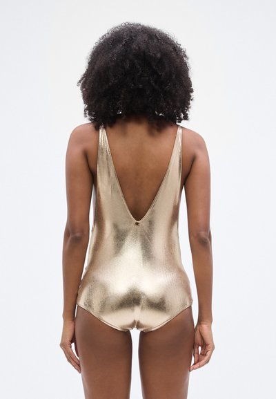 Metallist kuldse värvusega üheosaline ujumisriietus, millel on sügav V-kaelus tagaküljel, õhukesed rihmad ja sile tekstuur ning väike logoga detail alumise selja lähedal.