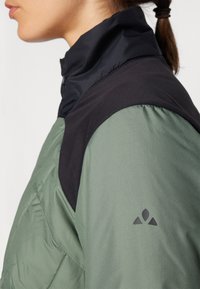 Veste verte et noire avec un col haut, un tissu texturé, un design d'épaule lisse et un logo sur la manche gauche.