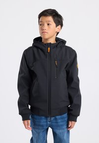 Musta pehmeäkuorinen takki hupulla, jossa on vetoketjullinen rintatasku ja joustavat hihansuut, yhdistettynä sinisiin farkkuihin.