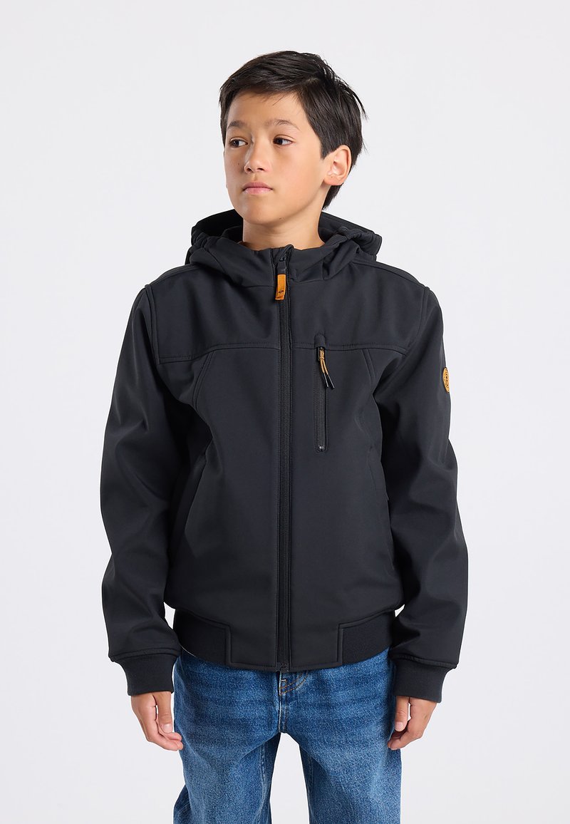 Musta pehmeäkuorinen takki hupulla, jossa on vetoketjullinen rintatasku ja joustavat hihansuut, yhdistettynä sinisiin farkkuihin.