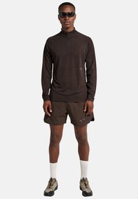 Mörkbrun långärmad zip-up-top, kombinerad med bruna shorts med en logotyp. Modellen har på sig solglasögon och sportiga skor. Vita strumpor synliga.