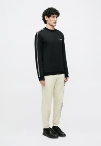 Juoda ilgomis rankovėmis džemperis su mažu logotipu ir kreminės spalvos sportinės kelnės su užrašu. Modelis dėvi juodus sportinius batelius.