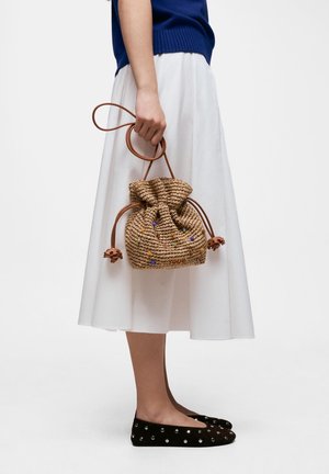 Mujer con falda midi blanca y zapatos planos negros sosteniendo un pequeño bolso marrón tejido con cordón y cuentas de colores.