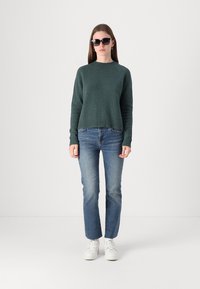 Pull en tricot vert avec des manches longues, un col rond et une finition côtelée ; associé à un jean bleu clair et des baskets blanches.