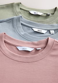 Trzy złożone koszulki z okrągłym dekoltem w stonowanym różu, jasnym niebieskim i oliwkowej zieleni z widocznymi metkami marki "NEXT."
