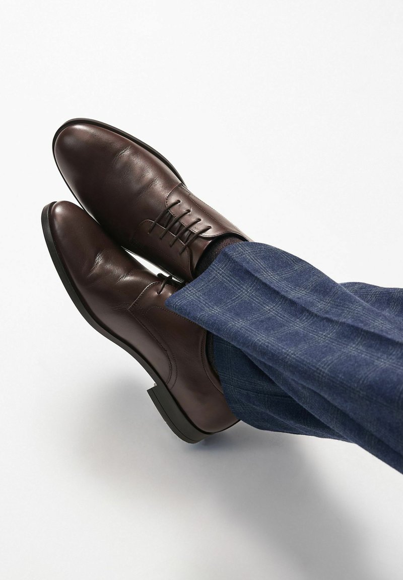 Zapatos de piel marrón con un acabado suave, con cordones y puntera redondeada, combinados con unos pantalones de cuadros azules a medida.
