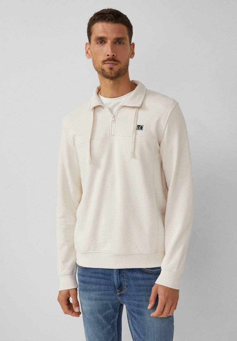 Sudadera de color beige claro con cuello de 1/4 de cremallera, cordones y un pequeño logo. Está hecha de un suave tejido texturizado. Mangas largas, corte relajado.