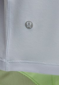 Close-up van lichtgrijze stof met een klein reflecterend Lululemon-logo boven groen gaas atletisch materiaal.