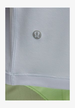 Zbliżenie na jasnoszary materiał z małym, odblaskowym logo Lululemon nad zielonym, siatkowym materiałem sportowym.