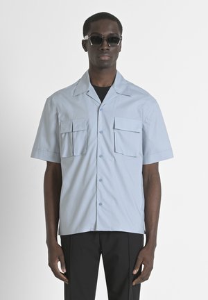 Lichtblauw kortemouwenshirt met twee voorzakken, puntige kraag en knopen in een relaxte pasvorm, gecombineerd met donkere broeken en een zonnebril.