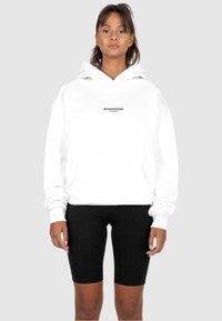 Weißer Hoodie mit einer Fronttasche, versehen mit einem schwarzen "METAMORPHOSIS"-Logo. Kombiniert mit schwarzen Radshorts aus glattem Stoff.