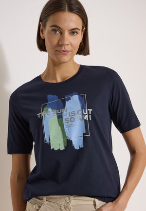 Femme aux cheveux courts et bruns portant un t-shirt bleu marine avec un motif de coups de pinceau bleus et verts et un texte imprimé, associé à un pantalon beige.