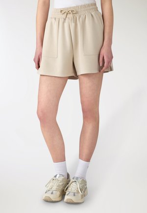 Persona che indossa pantaloncini beige con coulisse e tasche anteriori, calzini bianchi alti e sneakers beige con lacci, in piedi contro uno sfondo chiaro e uniforme.