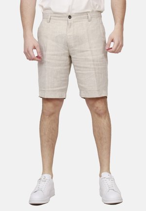 Uomo che indossa pantaloncini beige fino al ginocchio, camicia bianca e scarpe da ginnastica bianche, in piedi davanti a uno sfondo chiaro e uniforme.