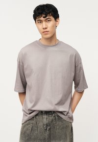 Νεαρός άνδρας με κοντά, σγουρά μαύρα μαλλιά, που φοράει ένα φαρδύ γκρι T-shirt και ψηλόμεσα ξεθωριασμένα μαύρα τζιν παντελόνια, στέκεται μπροστά σε ένα απλό φόντο.