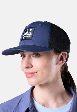 Mujer de piel clara y cabello oscuro recogido en una coleta, lleva una gorra azul y negra de Rossignol y una camisa azul clara, mirando ligeramente hacia la derecha.