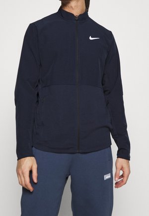 Osoba ubrana w czarną kurtkę na zamek z białym logo Nike na piersi i niebieskie spodnie z małą prostokątną metką.