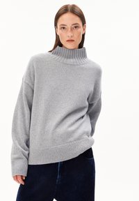 Jeune femme avec des cheveux bruns lisses portant un pull à col roulé en maille gris clair et un pantalon en velours côtelé bleu foncé, se tenant contre un fond blanc.