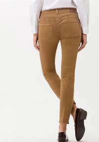 Jean skinny en denim beige avec une coupe ajustée, cinq poches, et un patch de marque sur la ceinture arrière. Texture lisse et design sur mesure.