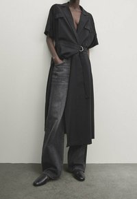 Person, der einen taillierten, kurzärmeligen schwarzen Mantel über lockeren dunkelgrauen Jeans und schwarzen spitzen Schuhen trägt, vor einer schlichten Wand.