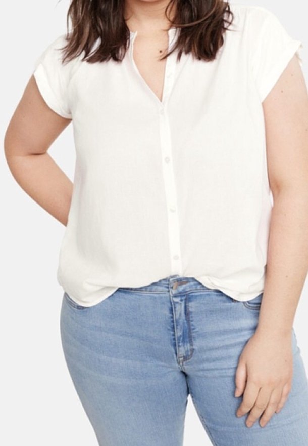 Vrouw draagt een witte blouse met korte mouwen en knopen, ingestopt in lichtblauwe spijkerbroek, staand met één hand op de heup.