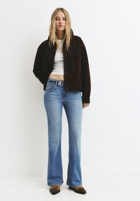 Rjava semiš jakna, skrajšana zasnova z gumbi, v kombinaciji s svetlo modrimi flared jeans. Obutev vključuje rjavo-črno barvno shemo.