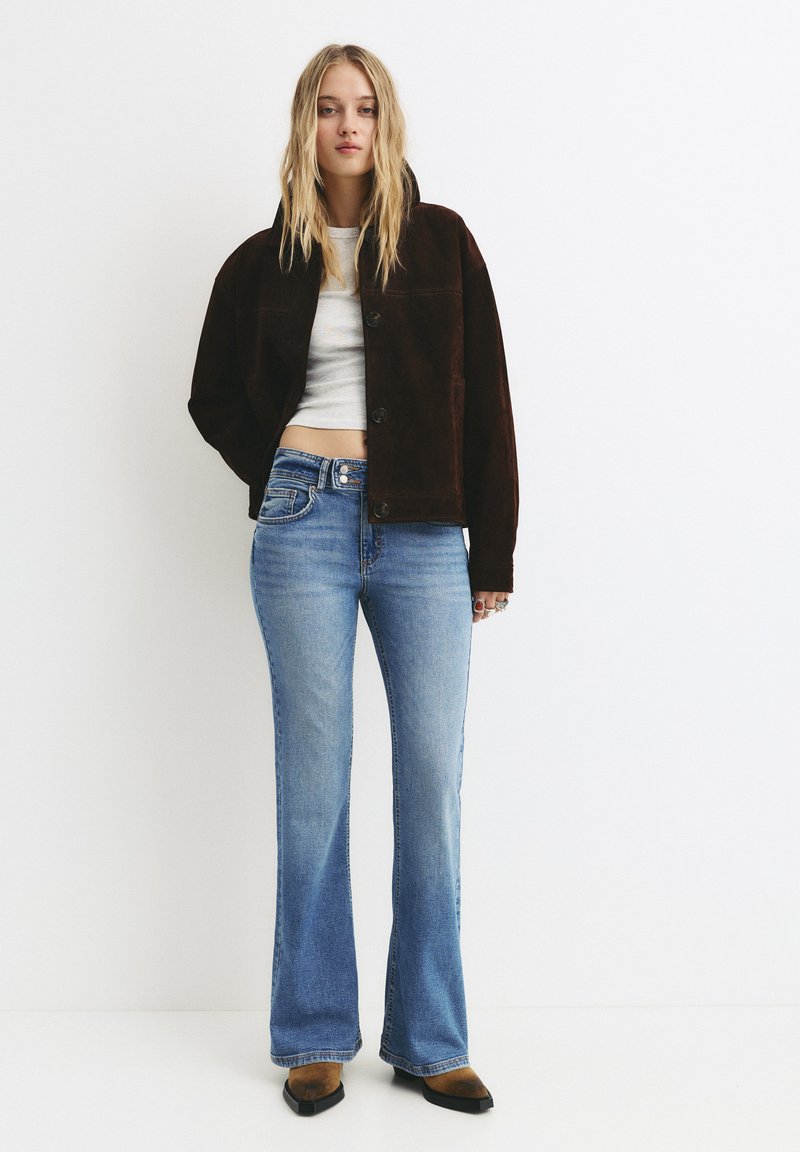 Rjava semiš jakna, skrajšana zasnova z gumbi, v kombinaciji s svetlo modrimi flared jeans. Obutev vključuje rjavo-črno barvno shemo.