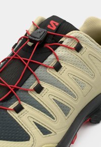 Zapatilla de running ligera con una parte superior de malla beige y negra, sistema de cordones rojo, paneles texturizados y suela de goma.