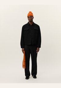 Chaqueta negra texturizada con dos bolsillos en el pecho, combinada con pantalones negros y un gorro de punto naranja brillante, sosteniendo una bufanda naranja con flecos.