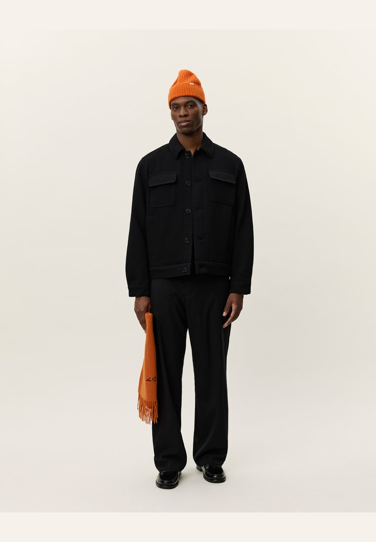 Chaqueta negra texturizada con dos bolsillos en el pecho, combinada con pantalones negros y un gorro de punto naranja brillante, sosteniendo una bufanda naranja con flecos.