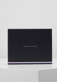 Svart rektangulær boks med en glatt tekstur, med en hvit logo "TOMMY HILFIGER" og en farget aksentstripe nederst.
