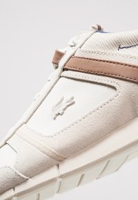 Sneaker beige chiaro realizzato in pelle e suede. Presenta una fascia marrone, dettagli testurizzati e un logo prominente sul lato. Suola in gomma.
