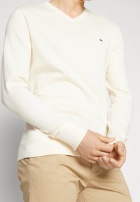 Maglione color crema a scollo a V in morbido tessuto lavorato a maglia, con polsini a costine e un piccolo dettaglio del logo sul petto. Abbinato a un paio di pantaloni beige.
